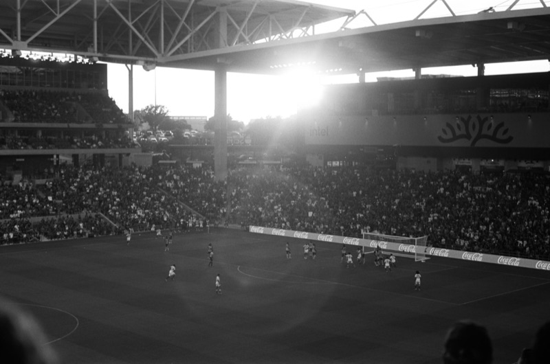 Black-and-white view of the Q2 Stadium field in Austin. Players on the pitch as sunlight flares through the stands